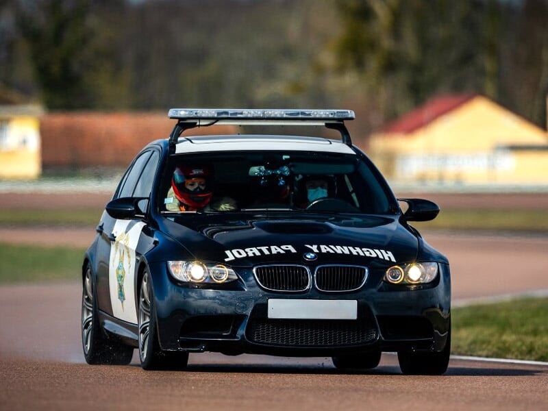 Voiture de police noire sur circuit, vue de face avec deux personnes à l'intérieur portant des casques.
