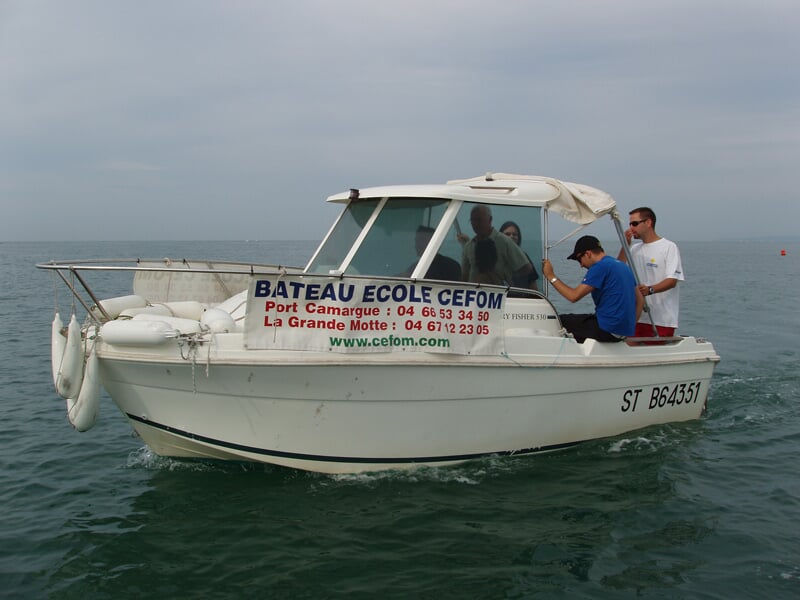 Petit bateau école CEFOM avec plusieurs personnes à bord naviguant sur une mer calme sous un ciel nuageux gris.