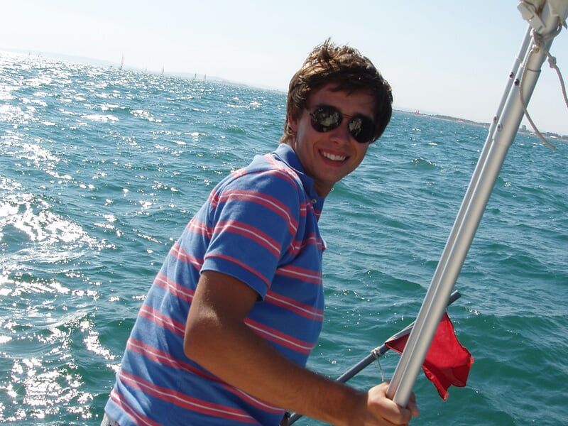 Jeune homme souriant en lunettes de soleil sur un bateau, tenant une barre avec la mer bleue en arrière-plan sous un ciel clair.