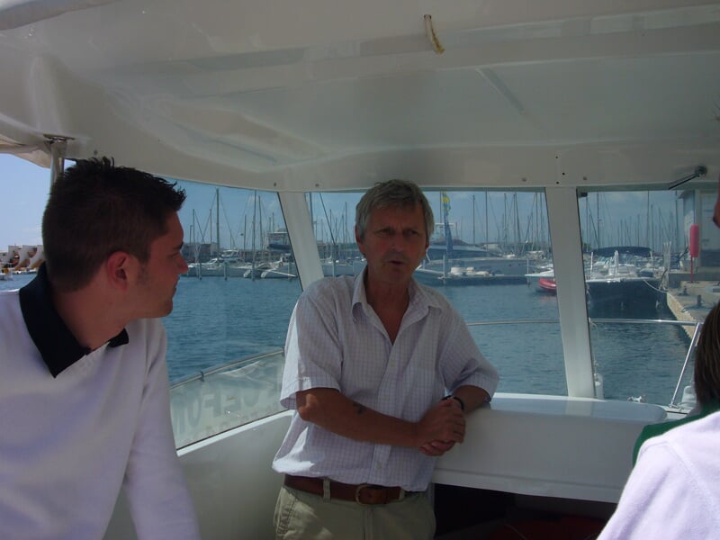 Trois hommes discutent à l'intérieur d'un bateau, avec une vue sur un port rempli de voiliers et un ciel dégagé.