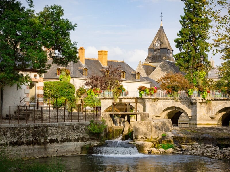 Village pittoresque avec un pont en pierre sur une rivière calme, maisons anciennes et clocher en arrière-plan.