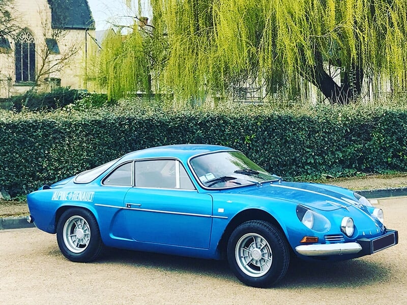 Voiture de sport bleue classique garée devant une haie et un grand saule pleureur en arrière-plan.
