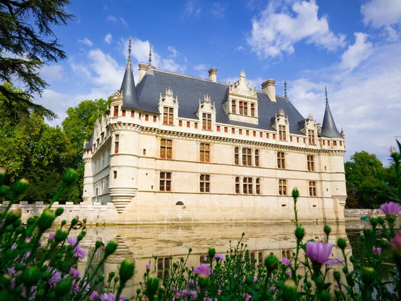 Château blanc majestueux entouré d'eau avec un ciel bleu et des fleurs violettes au premier plan.