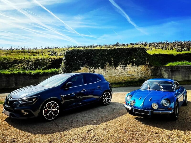 Deux voitures modernes et sportives garées sur un sol en gravier avec un mur de pierre et ciel bleu.