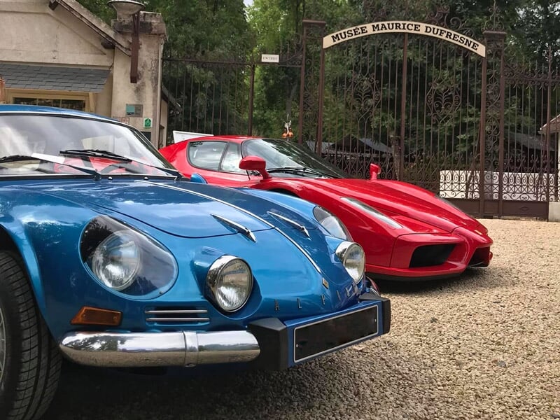 Voiture bleue classique et voiture rouge sportive garées devant un musée avec une grille en fer forgé.