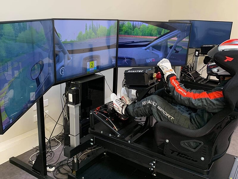  Un simulateur de course équipé de trois écrans incurvés, d'un volant Fanatec, de pédales et d'un siège baquet, avec un utilisateur portant une combinaison Renault Sport et un casque.