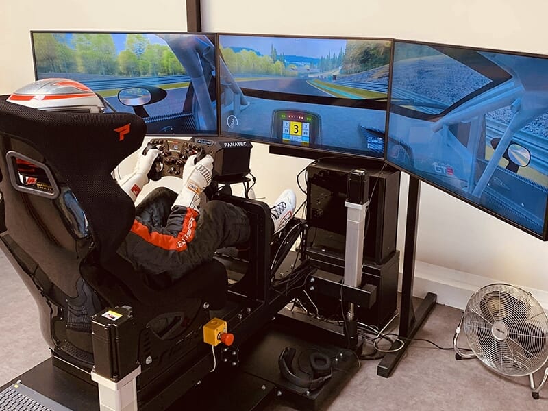 Vue arrière d'un utilisateur dans un simulateur de course avec trois écrans affichant une piste de course, des équipements Fanatec et un ventilateur à proximité.