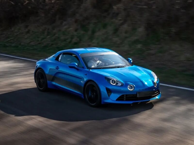Voiture de sport bleue en mouvement sur une route bordée d'arbres, avec un conducteur visible à l'intérieur.