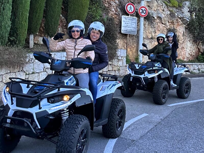 Groupe de personnes en quad sur une route avec des casques, prêt pour une balade, bordée par des murs en pierre et des panneaux de signalisation.