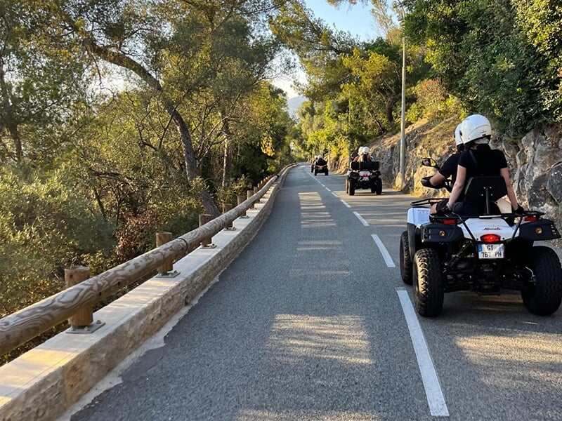 Vue arrière de quads roulant sur une route sinueuse entourée de végétation et de montagnes au loin.
