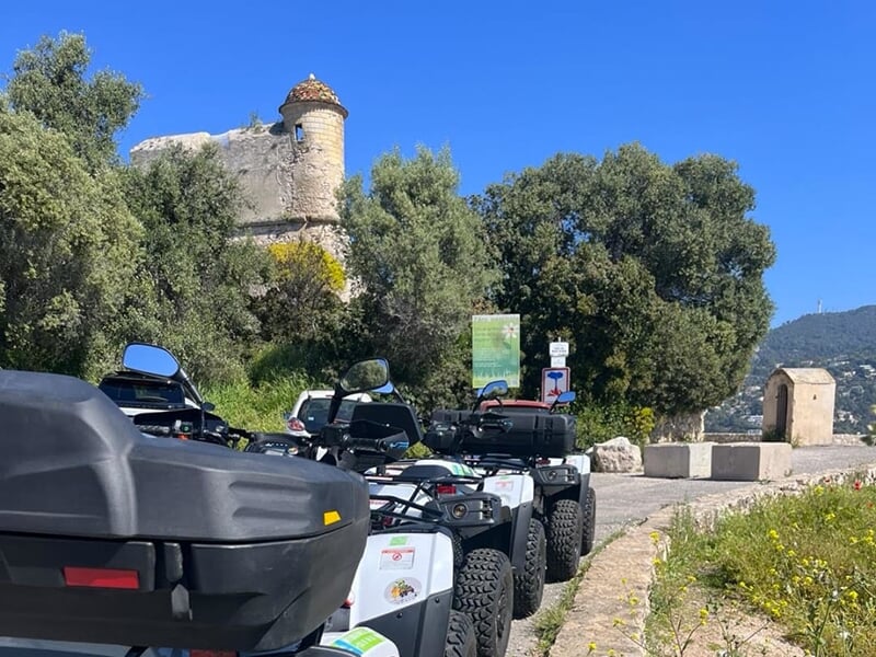 Plusieurs quads garés près d'une tour ancienne entourée de végétation sous un ciel bleu éclatant.