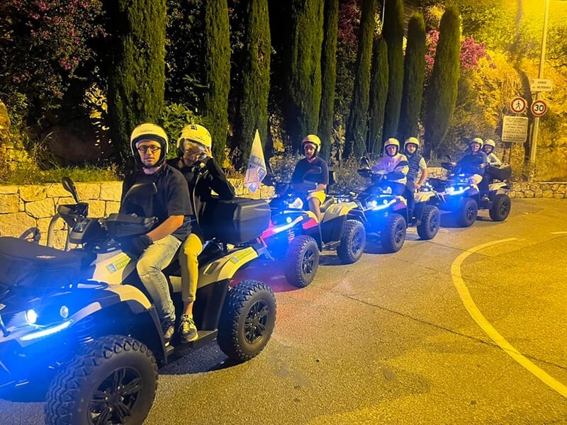 Groupe de quads en file, éclairés par des phares lumineux, roulant de nuit sur une route bordée de cyprès.