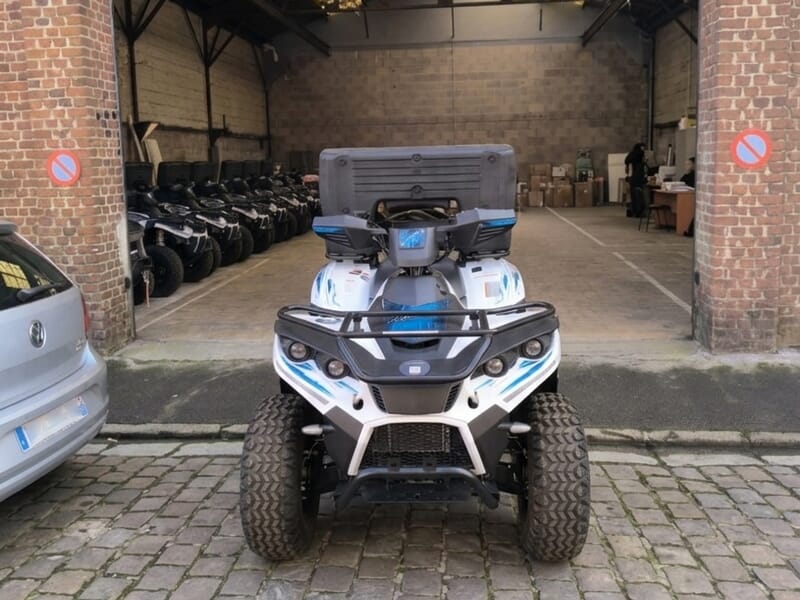 Quad blanc et bleu garé devant un garage avec plusieurs autres quads alignés à l'intérieur, sol en pavés.