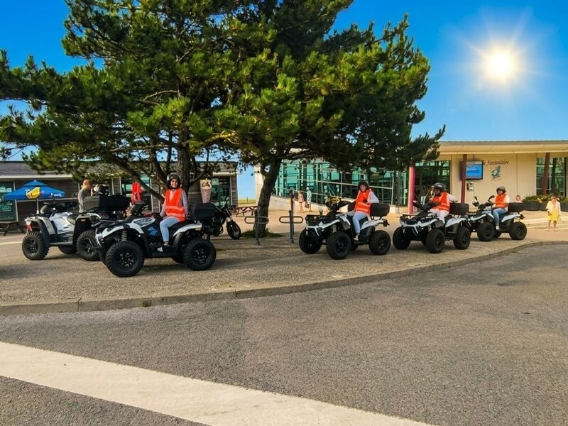 Groupe de personnes en gilets orange assises sur des quads alignés sur un trottoir avec un ciel bleu et un soleil brillant.
