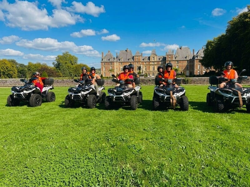 Cinq personnes en gilets orange posant sur des quads sur une pelouse verte devant un grand château sous un ciel bleu.