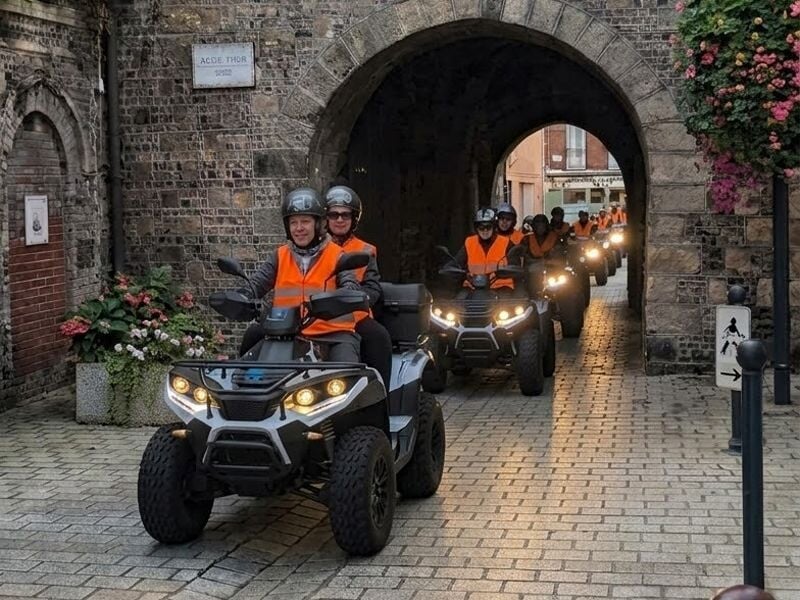Groupe de personnes en gilets orange conduisant des quads en file indienne sous une arche en pierre dans une rue pavée.