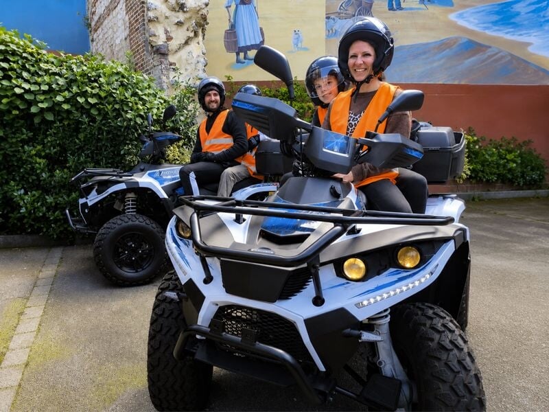 Trois personnes souriantes en gilets orange assises sur des quads dans une cour avec des murs peints et des plantes.