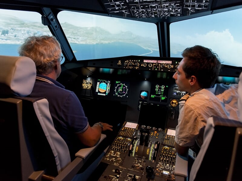 Deux personnes dans un simulateur de pilotage d'avion, observant les instruments et l'écran de contrôle devant eux.
