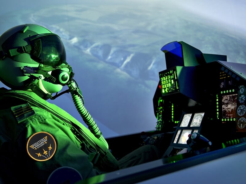Pilote en combinaison et casque dans un simulateur de vol militaire, avec vue sur les écrans et le paysage extérieur.