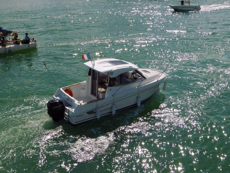 Petit bateau blanc naviguant sur une mer calme avec un drapeau français à l'arrière sous un ciel ensoleillé.