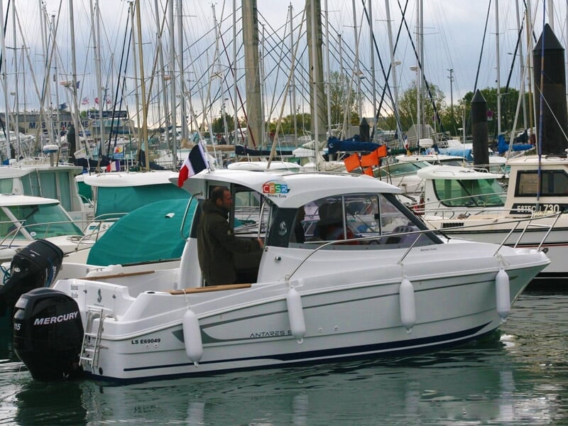 Bateau blanc amarré dans un port avec plusieurs voiliers en arrière-plan et un homme debout à bord.
