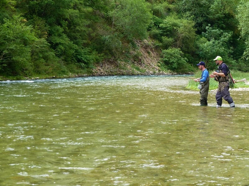 Deux pêcheurs en waders pêchent dans une rivière entourée d'arbres verts et d'une nature luxuriante.