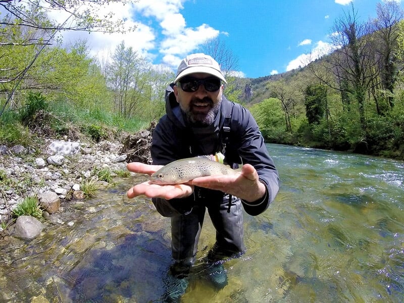 Un homme souriant montre une truite qu'il tient dans ses mains au bord d'une rivière ensoleillée.