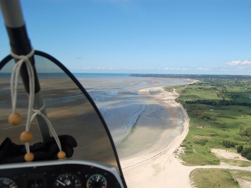 Cockpit d’ULM en approche d’une plage sinueuse à marée basse, avec prairies verdoyantes et ciel bleu en arrière-plan.