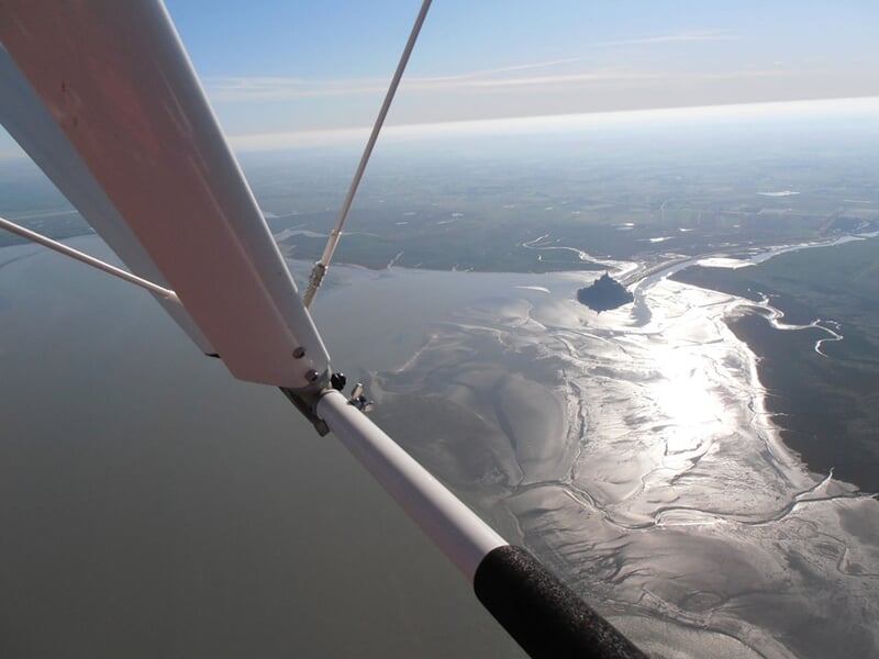 Vue aérienne d’un estuaire scintillant à marée basse avec le Mont-Saint-Michel à l’horizon sous un ciel dégagé.