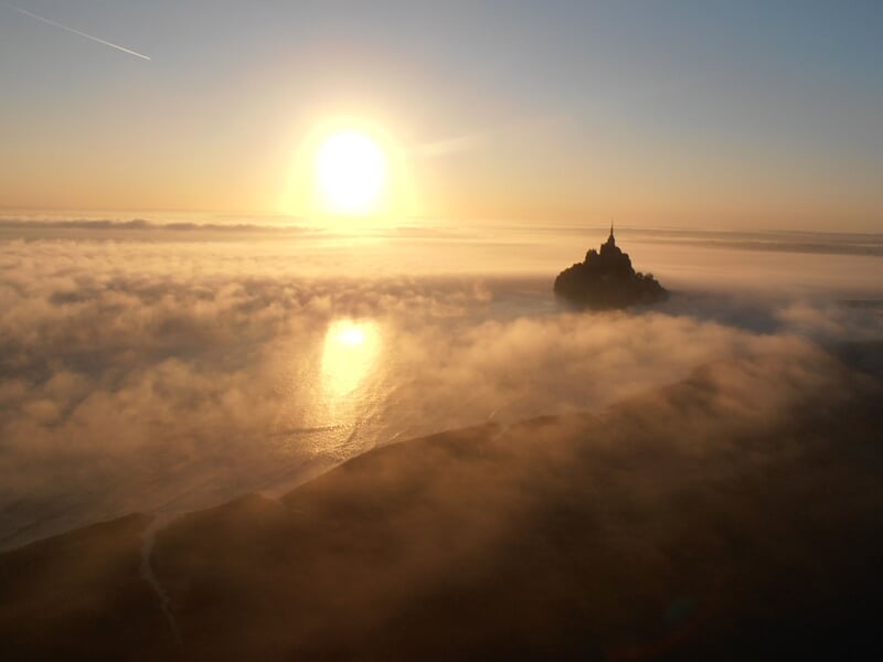Vue aérienne du Mont-Saint-Michel au coucher du soleil, avec des nuages bas et une lumière dorée.