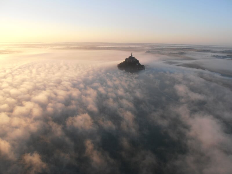 Silhouette du Mont-Saint-Michel émergeant d’une mer de nuages au lever du soleil, baignée d’une lumière dorée.