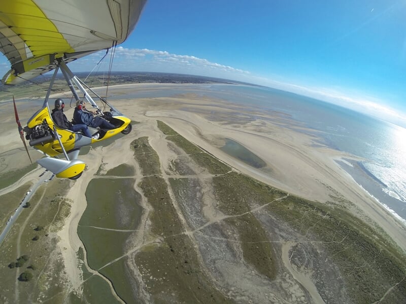 ULM jaune survolant un vaste espace de dunes, zones humides et plages à marée basse sous un soleil éclatant.