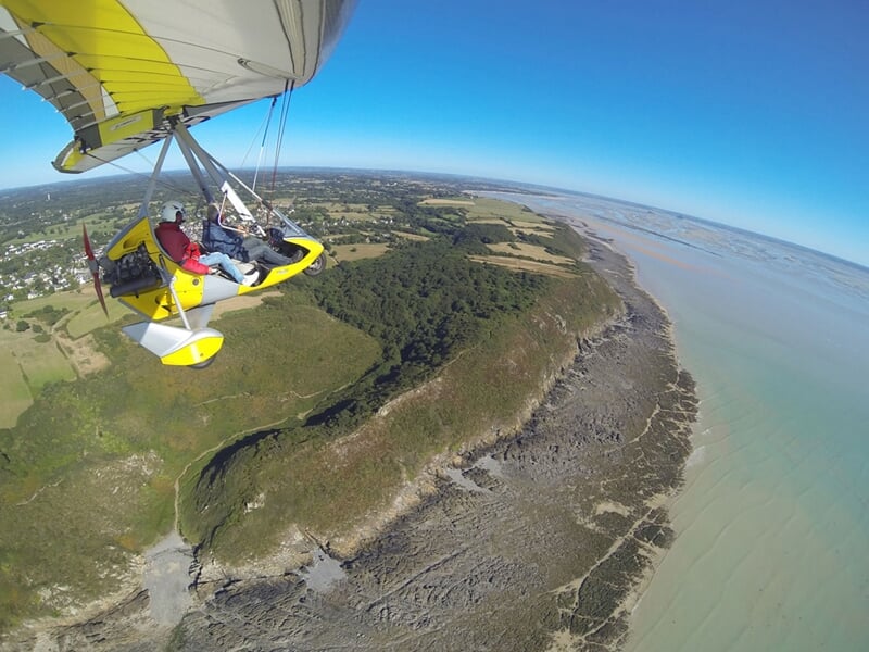 ULM jaune en vol longeant une falaise verdoyante bordée par une plage rocheuse et une mer turquoise sous ciel bleu.