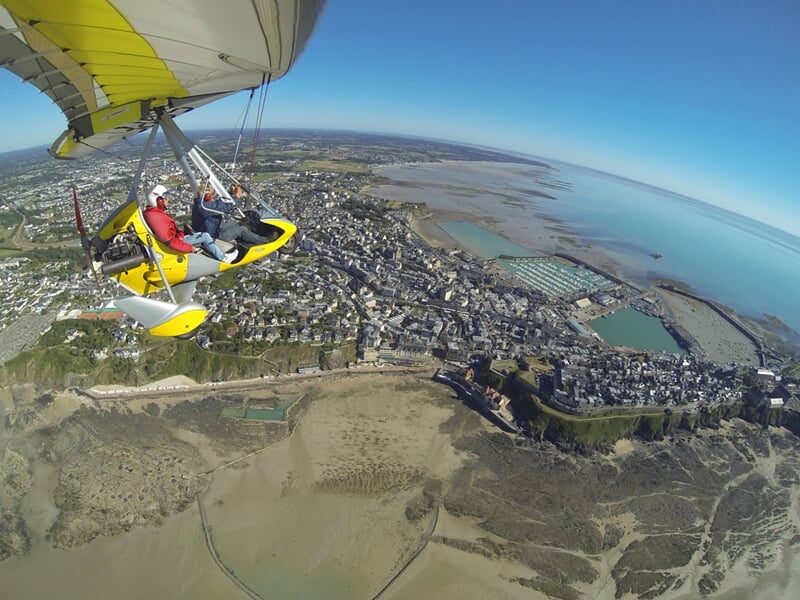 ULM jaune survolant une ville côtière avec plages, bassins ostréicoles et étendue d'eau turquoise.