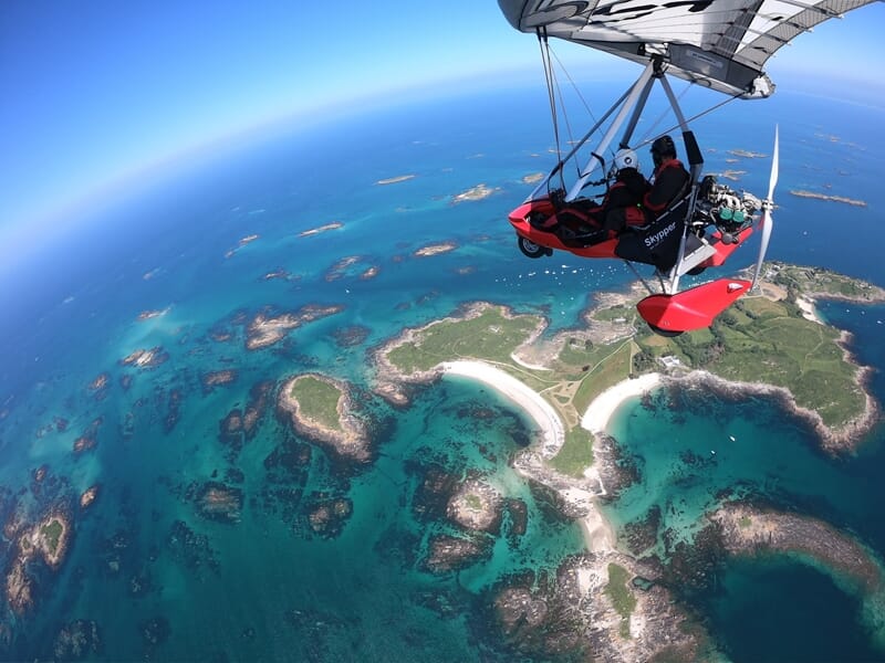ULM rouge survolant une côte découpée avec plages blanches, eau turquoise et îlots rocheux sous un ciel bleu dégagé.