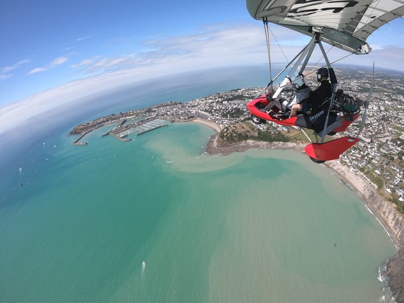 ULM rouge survolant un port de plaisance en bord de ville avec plage, falaises et eau vert turquoise.