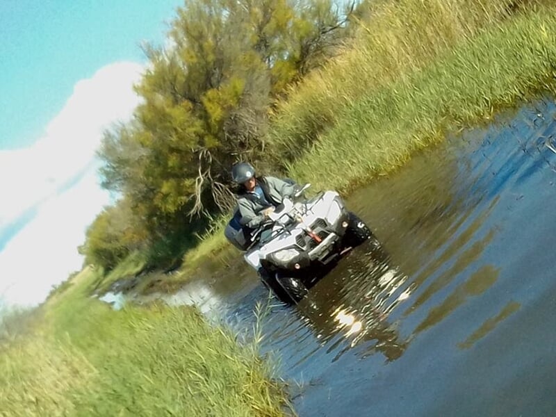 Personne en casque conduisant un quad dans un chemin d'eau entouré de végétation verte dense sous un ciel bleu.