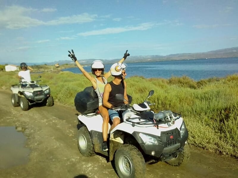 Deux personnes souriantes sur un quad blanc sur un chemin près d'un lac, avec des collines en arrière-plan.