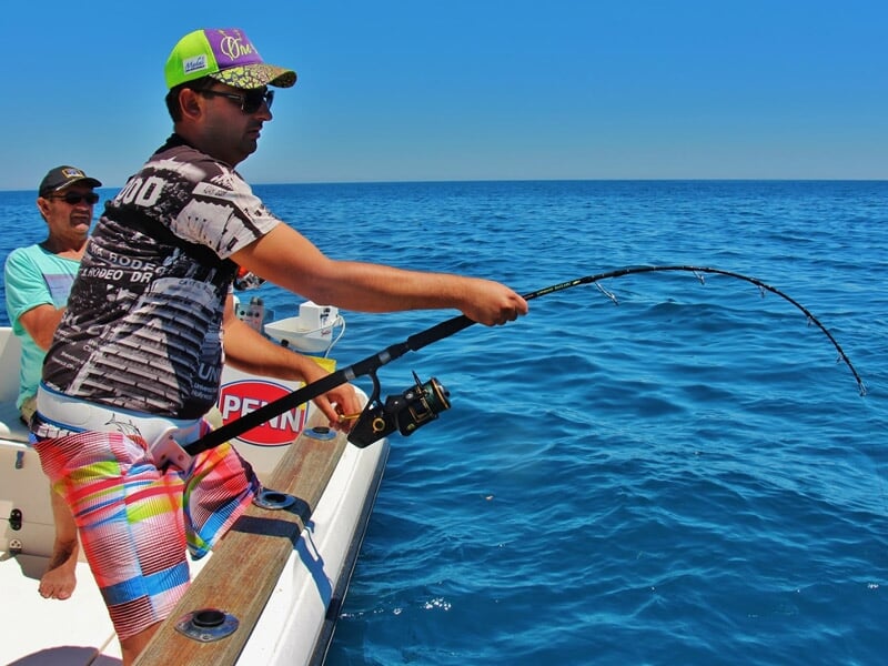 Homme en short coloré et casquette pêche au gros sur un bateau, canne à pêche pliée sous le soleil bleu.