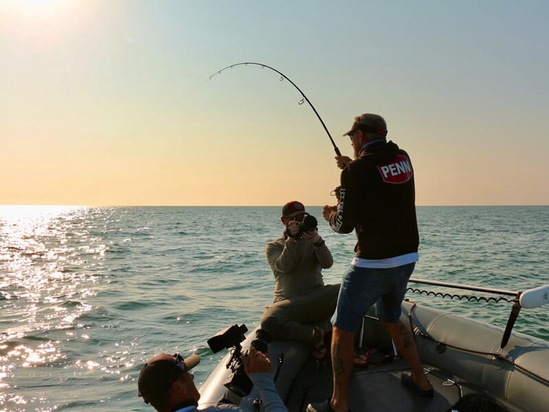 Pêcheur debout sur un bateau au coucher du soleil, tenant une canne à pêche pliée, entouré de photographes.