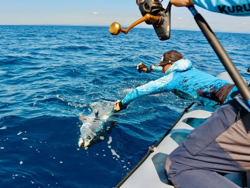 Pêcheur en tenue bleue attrape un gros poisson à la main depuis un bateau en pleine mer sous un ciel clair.