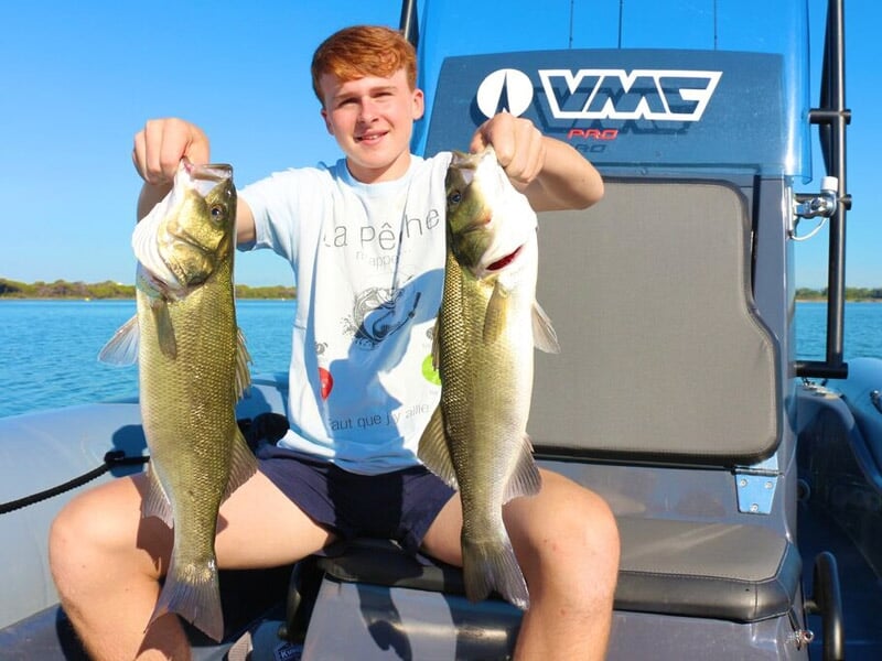 Jeune garçon assis sur un bateau montre fièrement deux gros poissons qu'il vient de pêcher, ciel clair.
