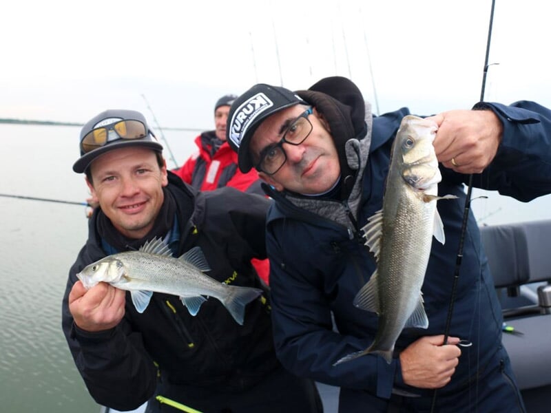 Deux hommes souriants montrent leurs poissons fraîchement pêchés sur un bateau, ambiance conviviale.