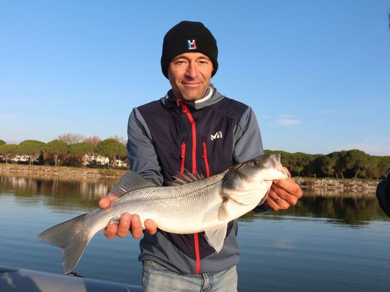Homme en veste et bonnet tient un gros poisson blanc sur un bateau, paysage naturel en arrière-plan.