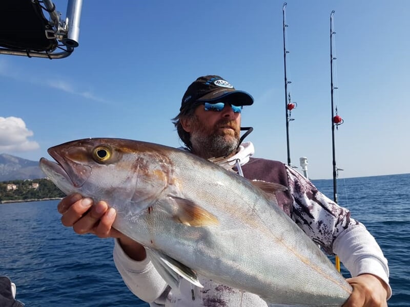 Pêcheur avec casquette et lunettes tenant un grand poisson argenté sur un bateau en mer.