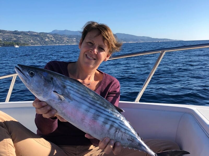 Femme souriante tenant un poisson rayé sur un bateau avec la mer et les montagnes en arrière-plan.