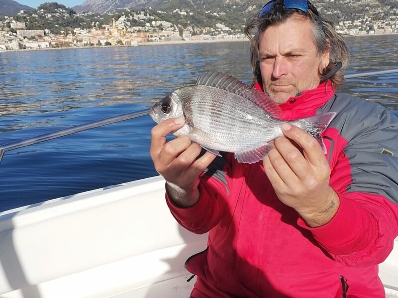 Homme en veste rouge tenant un petit poisson argenté sur un bateau en mer, paysage côtier visible.