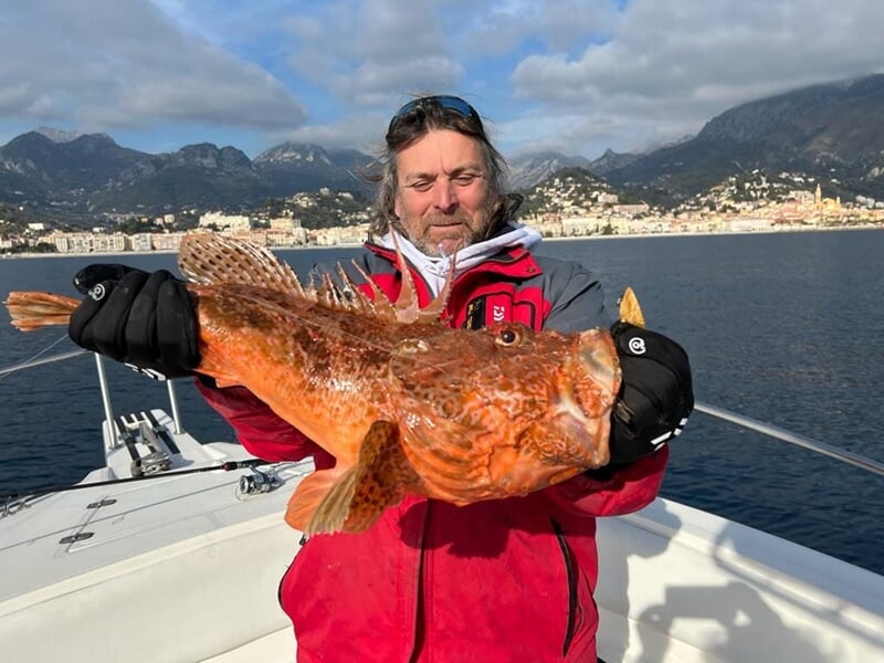 Homme en veste rouge tenant un gros poisson orange avec la ville et la mer en arrière-plan.