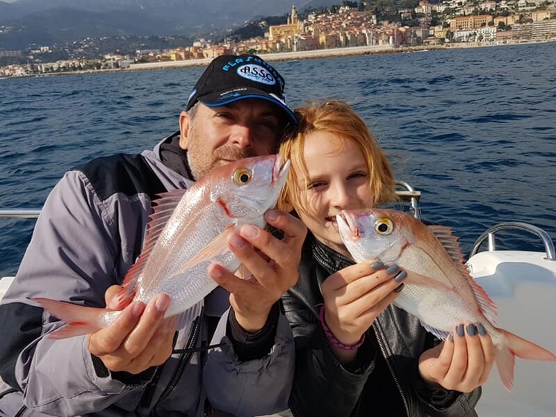 Deux personnes souriantes tenant chacune un petit poisson rose sur un bateau en mer à Monaco.