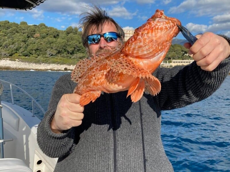 Homme avec lunettes de soleil tenant un gros poisson orange sur un bateau près de la côte.